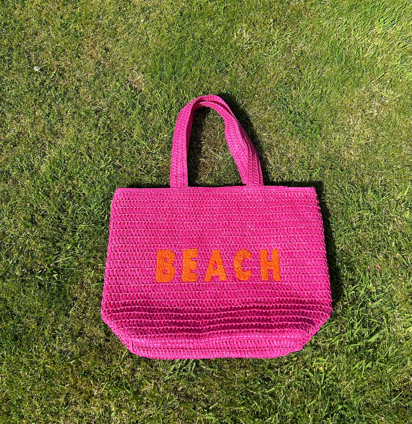 A pink beach bag with the word 'BEACH' written in large orange letters on a grassy surface.