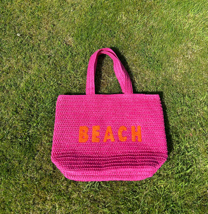 A pink beach bag with the word 'BEACH' written in large orange letters on a grassy surface.