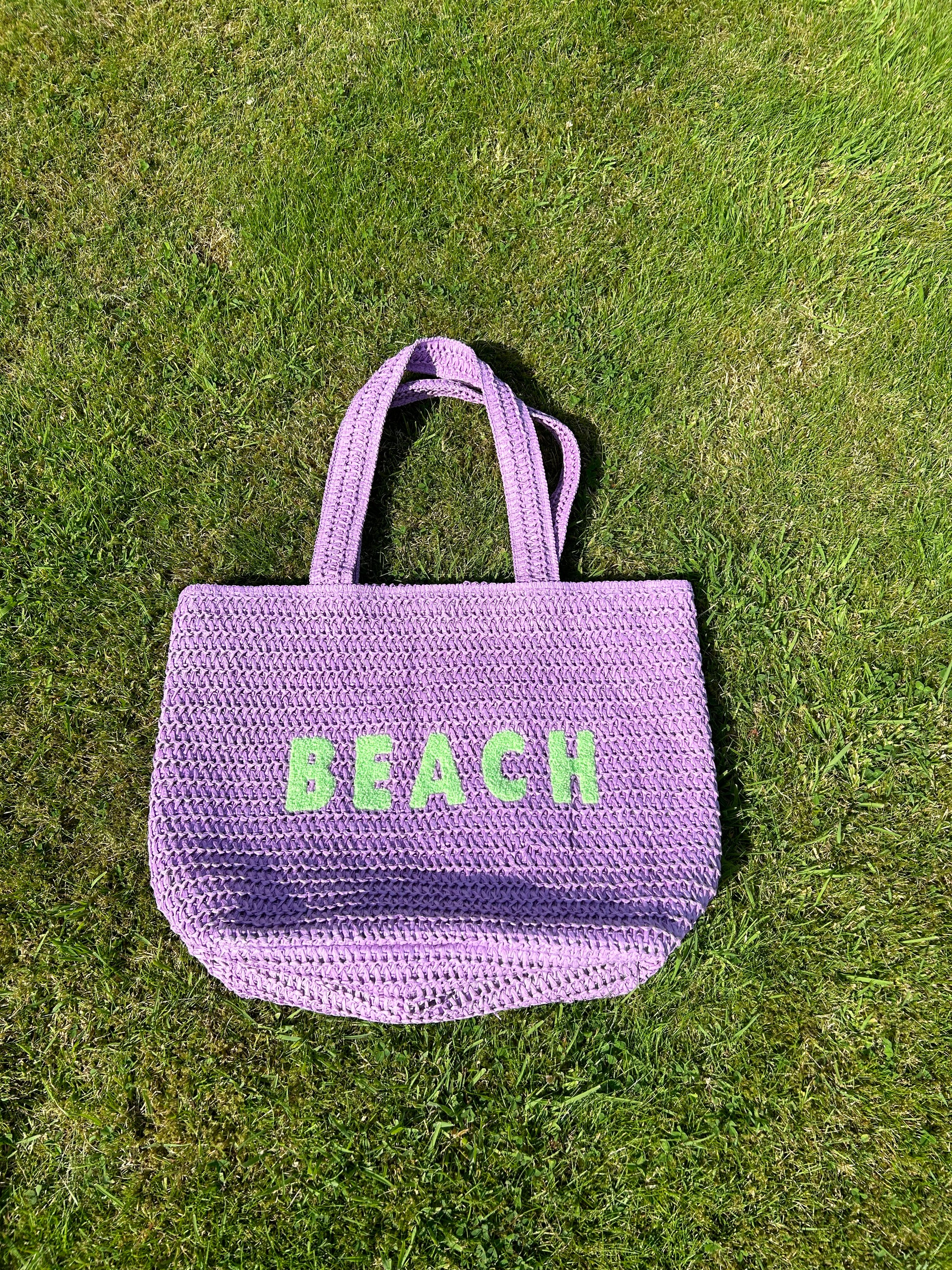 A purple beach bag with the word 'BEACH' written in large green letters on a grassy surface.